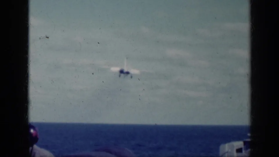 The aircraft appears smaller as it flies above the ocean, with part of a deck observer visible at the edge of the 8mm frame.