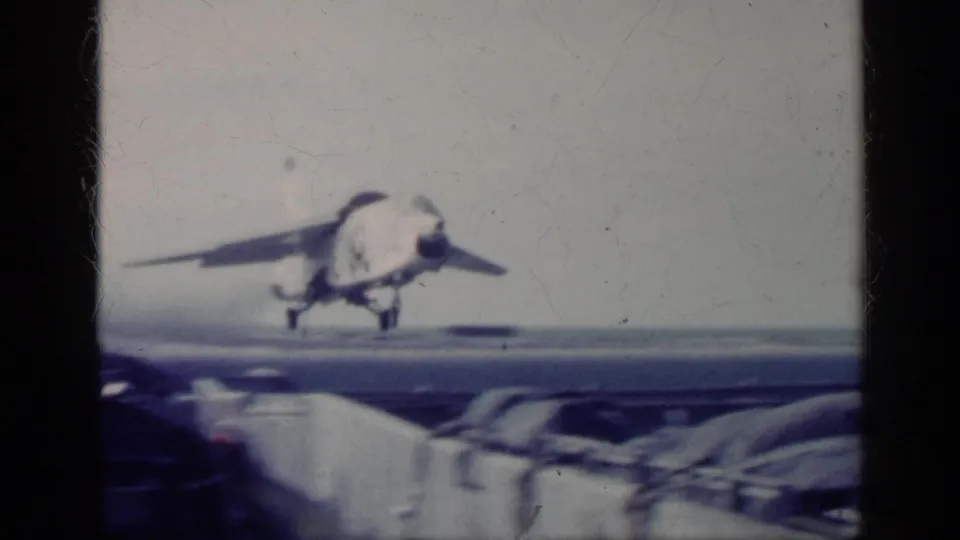 1962 Cuba-associated 8mm frame showing a Navy jet just above the carrier deck with ocean and overcast sky behind it.
