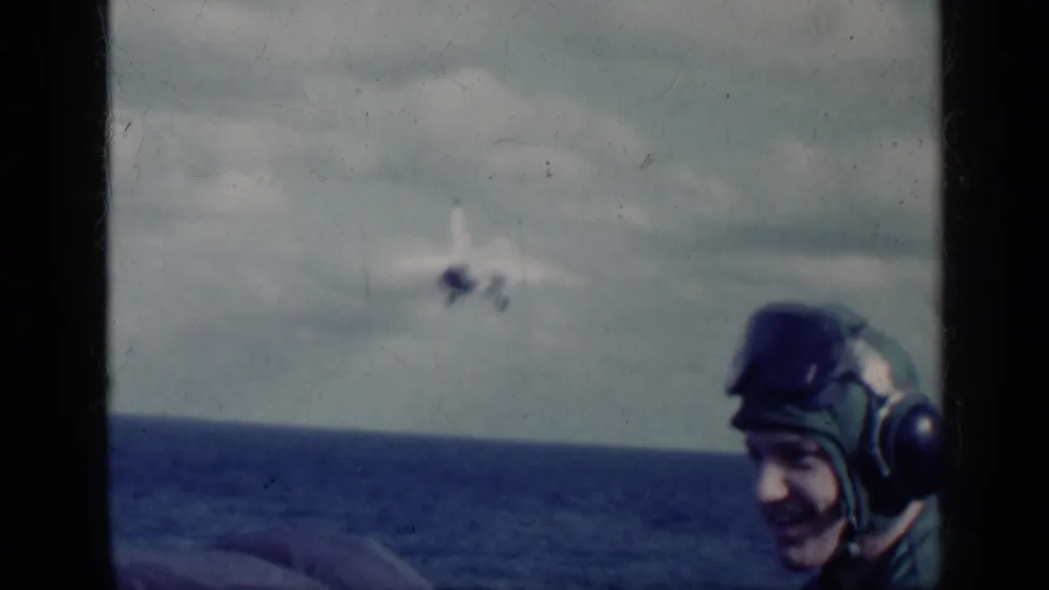 1962 Cuba-associated 8mm frame showing a helmeted Navy crew member in the foreground as an aircraft flies over the sea.