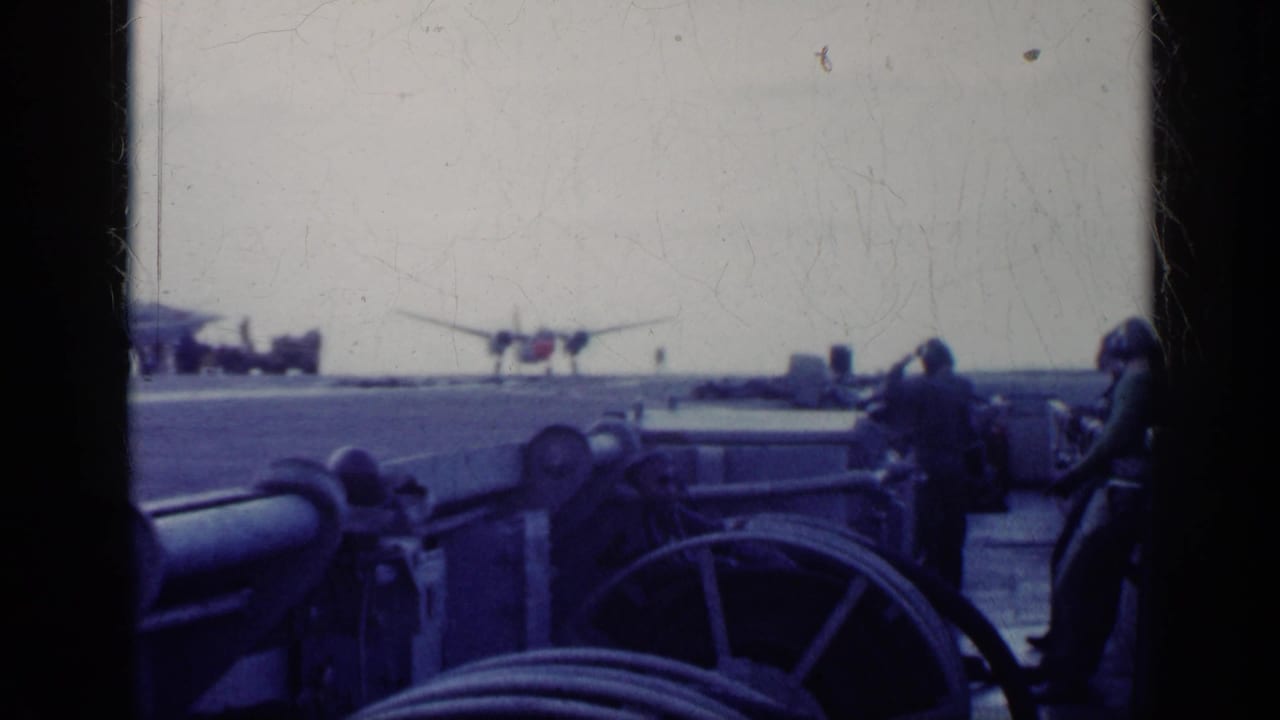Carrier deck seen through dark framing with crew and a distant twin-engine aircraft.
