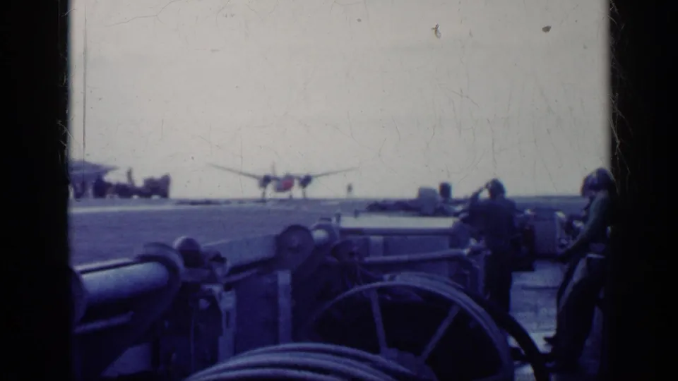 1962 Cuba 8mm frame showing a ship deck, uniformed personnel, deck equipment, and a distant propeller aircraft under an overcast sky.