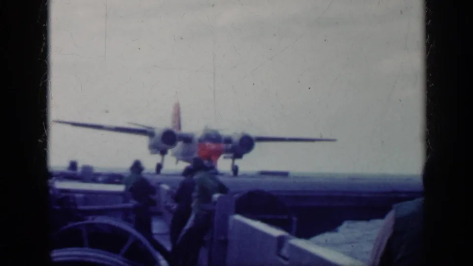 1962 Cuba 8mm frame showing a twin-engine propeller aircraft moving along a ship deck with uniformed personnel and deck gear in view.