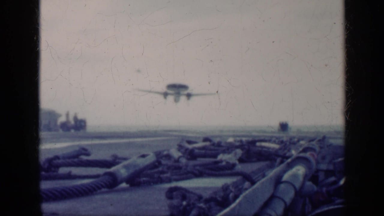 Propeller aircraft approaching a carrier deck over ropes and deck equipment.