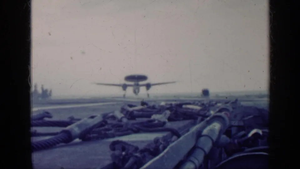 A propeller aircraft nears the flight deck in blue-tinted 8mm footage, seen past thick ropes and equipment on the carrier.