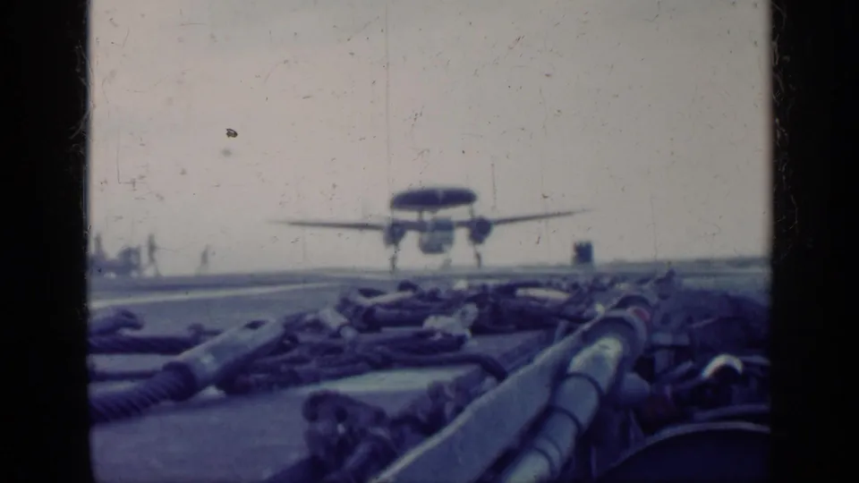 The aircraft is just above the carrier deck, filmed from a low deck-level angle with ropes and equipment in the foreground.