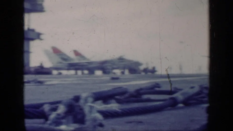 Carrier deck view after the landing pass, with heavy ropes in front, parked aircraft in the distance, and small personnel figures visible.