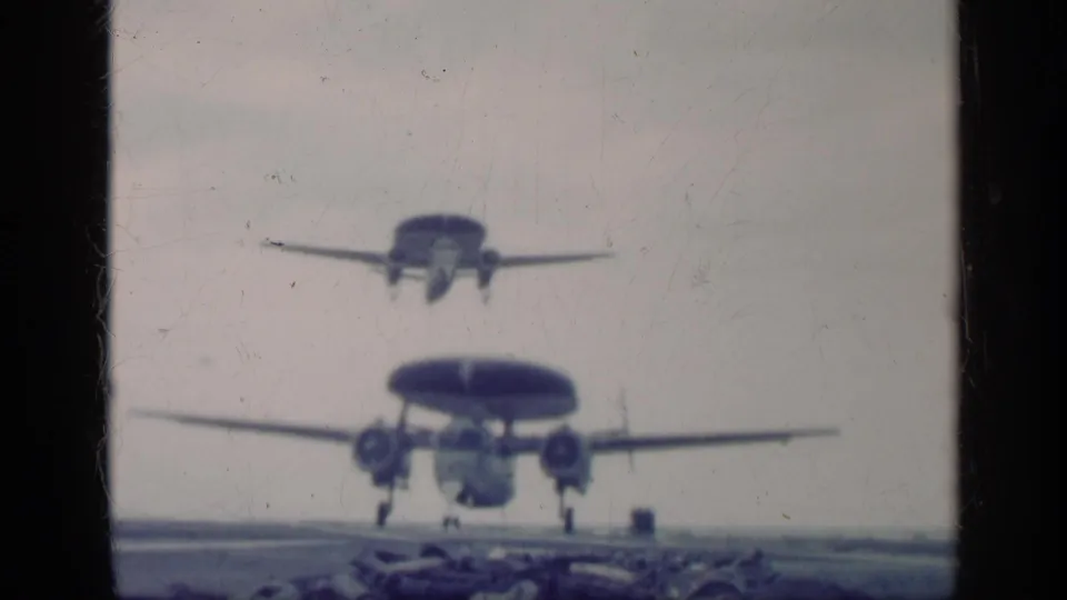 Blue-tinted 8mm frame of two radar-equipped military aircraft approaching above an aircraft carrier deck during the Cuba-labeled 1962 clip.