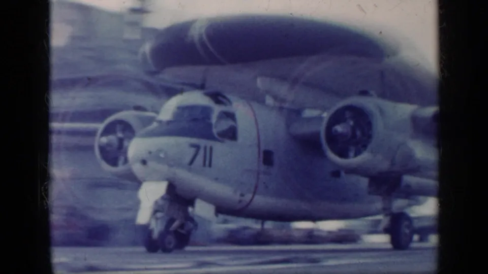 Close 8mm frame of a radar aircraft on an aircraft carrier deck, showing nose number 711, propeller engine, landing gear, and radar dome.