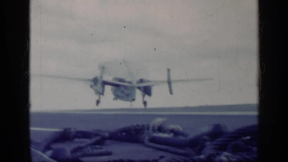 The U.S. Navy aircraft begins takeoff from the carrier deck, with deck equipment in the foreground and ocean horizon beyond.