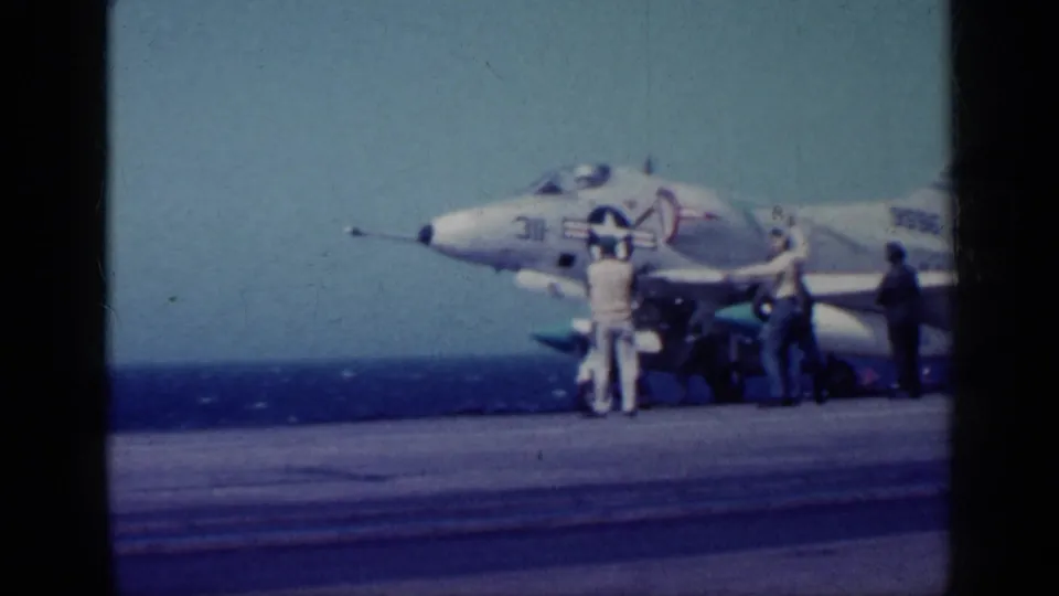 1962 Cuba-context frame showing a U.S. Navy jet on a carrier deck as several crew members stand near the nose and landing gear.