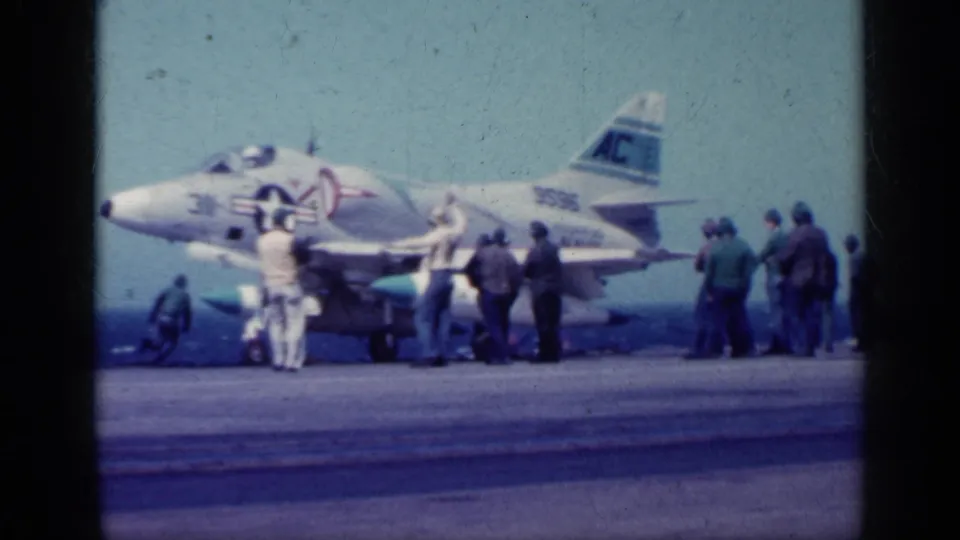 1962 Cuba-context frame showing a side view of a marked U.S. Navy jet while crew members stand around the aircraft on deck.