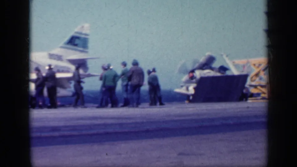 1962 Cuba-context frame showing crew on an aircraft carrier deck near a jet tail section and deck equipment, with ocean behind them.