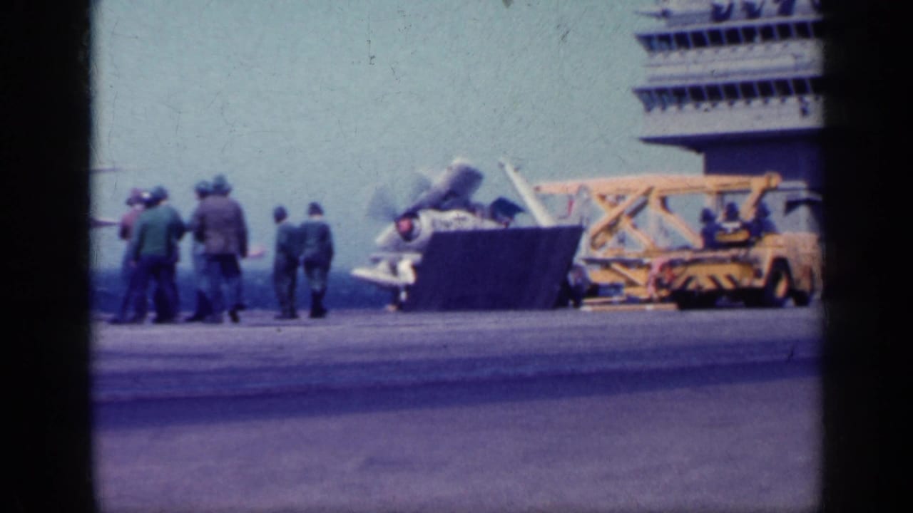 Carrier deck crew near military jets and a yellow tow vehicle with ocean beyond.