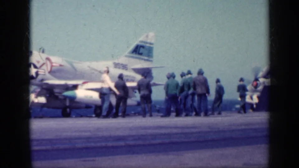 A carrier-deck jet with visible tail markings is surrounded by personnel during apparent launch or handling preparation.