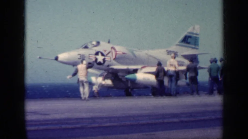 1960s 8mm frame of an A-4-type jet on a carrier deck as crew members cluster around the aircraft.
