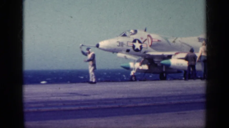 Carrier-deck view of a marked jet and nearby crew, including one person standing forward of the aircraft with arms raised.