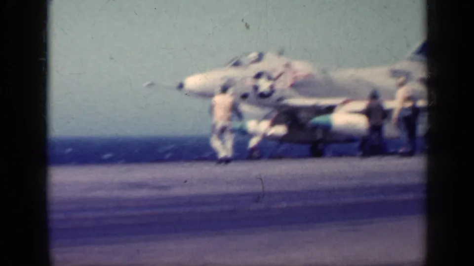 A soft 8mm frame shows a U.S. Navy jet marked with insignia on the carrier deck, with crew clustered near the plane and sea beyond.