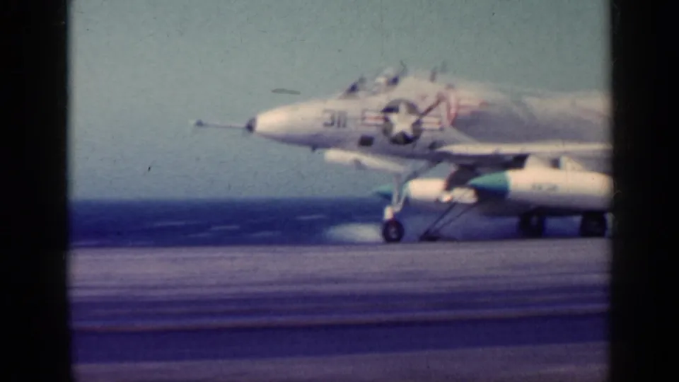 The jet appears larger in frame as it moves on the carrier deck, with Navy markings, landing gear, and ocean visible behind it.