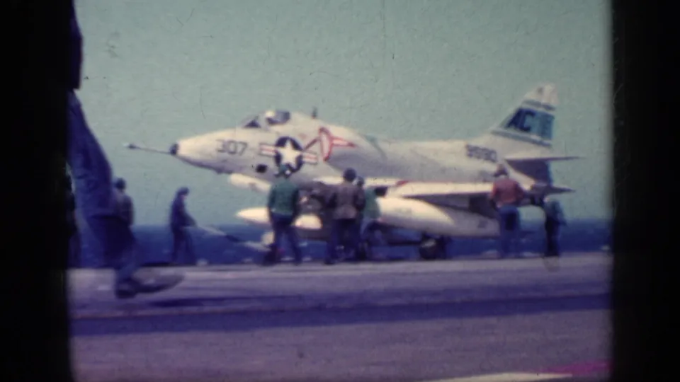 A U.S. Navy jet with visible markings sits on the carrier deck as crew gather and a figure passes close to the camera.