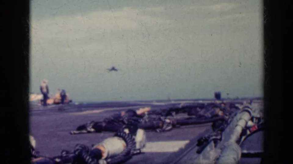 Carrier deck view with deck equipment in the foreground, crew at left, and a small jet rising beyond the runway against the horizon.