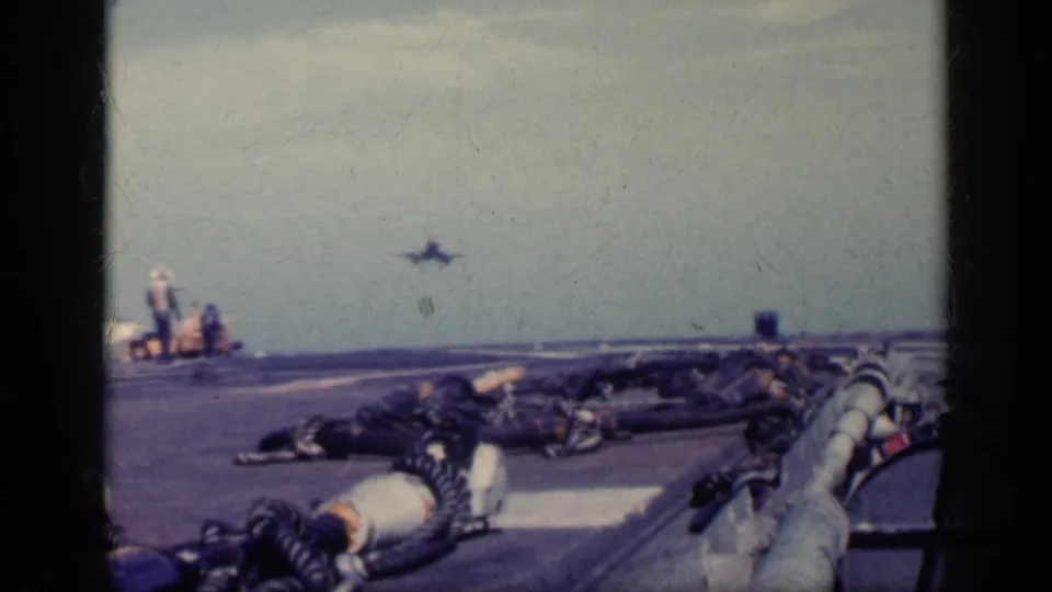 Soft 8mm frame of a distant military jet above the carrier flight deck, with crew figures and deck gear visible near the runway edge.