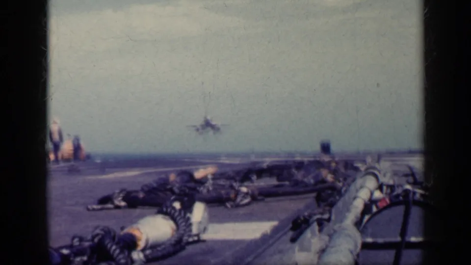 Faded 8mm carrier-deck frame showing a military jet near the horizon line, with deck equipment and the flight deck stretching toward the sea.