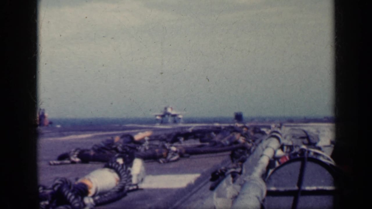 1962 Cuba metadata supports this view of a military jet nearing a carrier deck, seen past hoses, deck gear, ocean, and hazy sky.