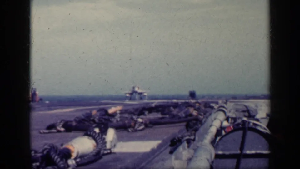 1962 Cuba metadata supports this view of a military jet nearing a carrier deck, seen past hoses, deck gear, ocean, and hazy sky.