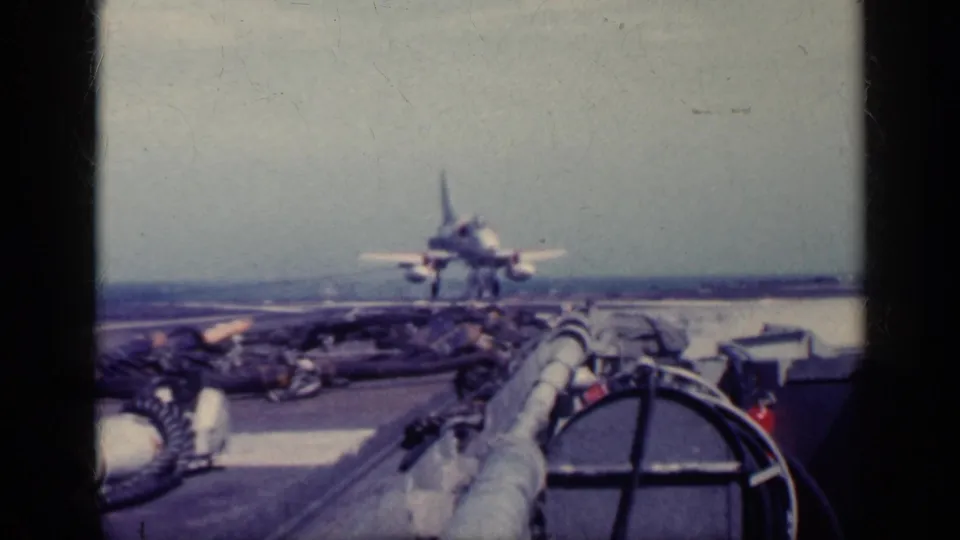 A military jet appears close to landing on an aircraft carrier flight deck, with deck equipment and the ocean horizon visible in the 8mm frame.