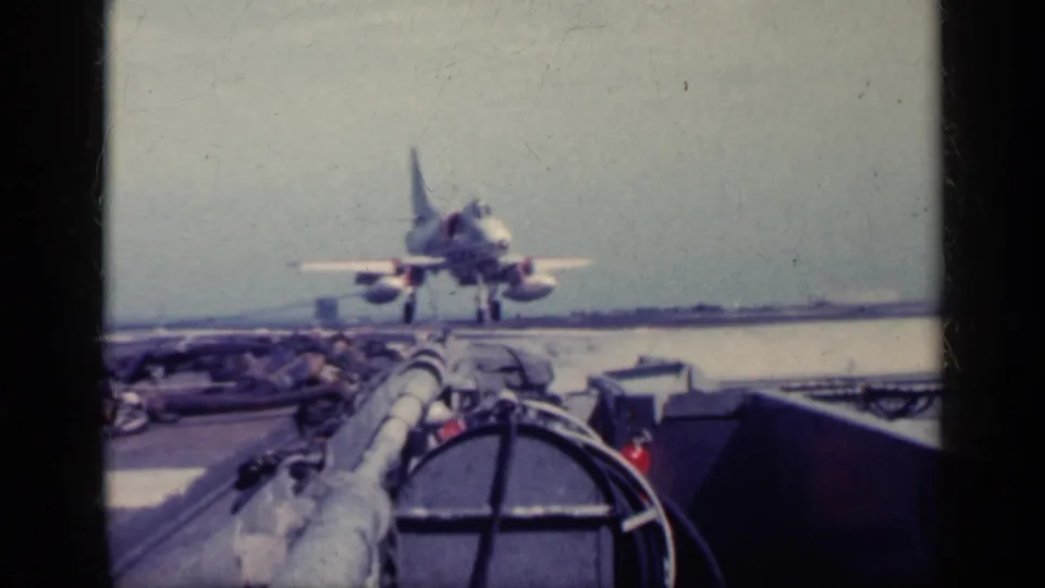 The jet is low over or on the carrier deck, with flight-deck hoses, equipment, sea, and hazy sky visible in the faded 8mm footage.