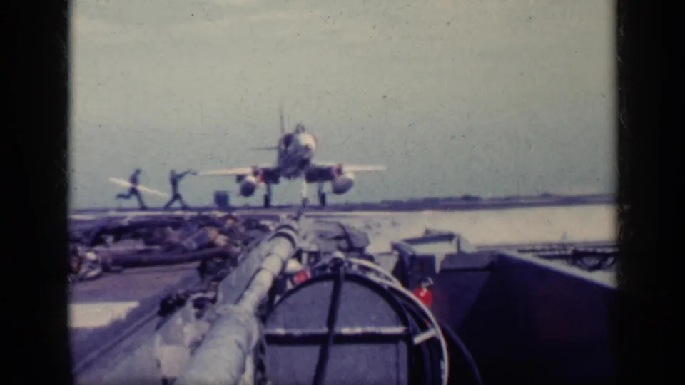 Deck personnel run toward a military jet on the aircraft carrier deck, with sea, equipment, and film gate edges visible around the frame.