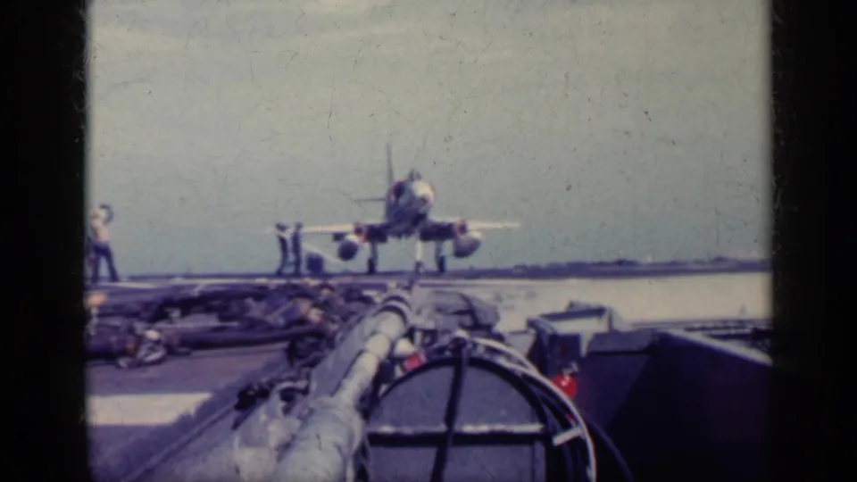 1962 Cuba metadata-supported 8mm frame showing a military jet on a carrier deck, viewed from a low angle with crew and sea visible.