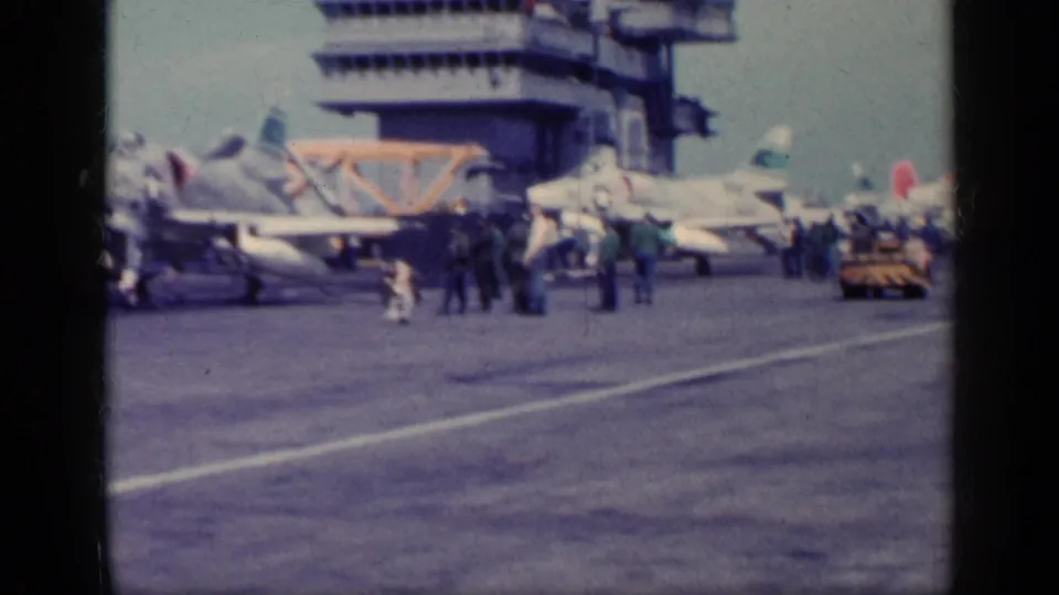 Carrier flight deck scene with parked military aircraft, crew figures, and the ship island structure in soft 8mm color footage.