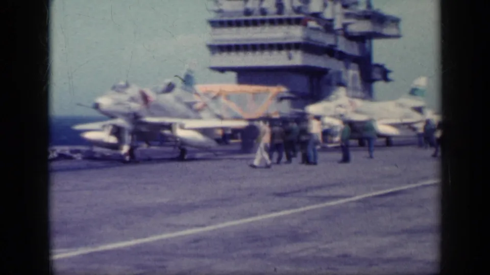 1960s carrier deck view showing military jets, crew figures, runway markings, and the island structure in faded 8mm footage.