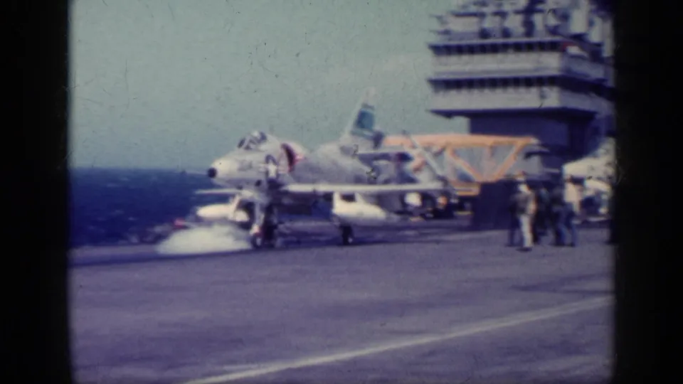A military jet appears on the aircraft carrier flight deck near crew members, with open water and the carrier island behind it.