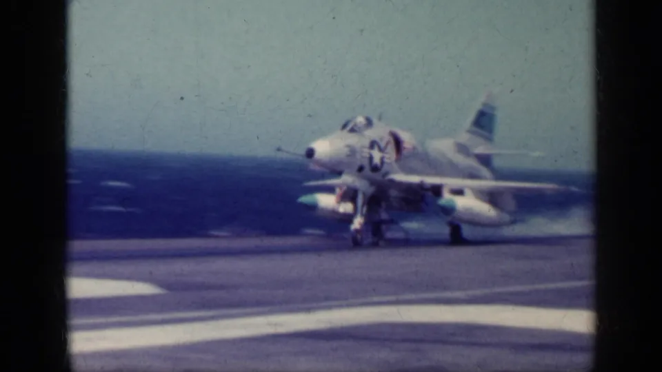 The carrier-deck view shows the Navy jet continuing its launch run, with landing gear down and the sea visible beyond the deck.