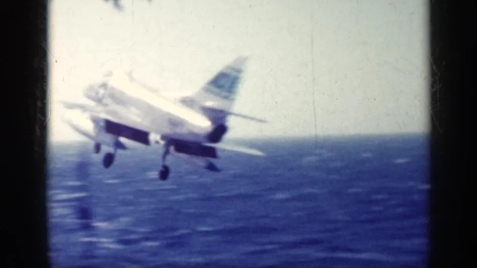 A U.S. Navy jet lifts from the carrier deck over the ocean, its landing gear still extended in grainy 8mm footage.