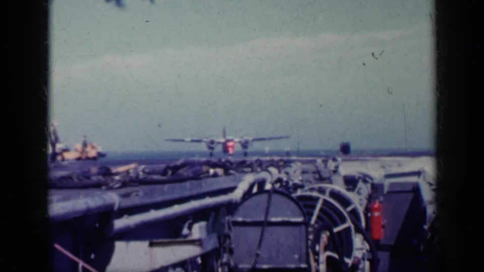 1962 Cuba 8mm frame with a military aircraft over or near an aircraft carrier deck, viewed from behind deck railings and equipment.