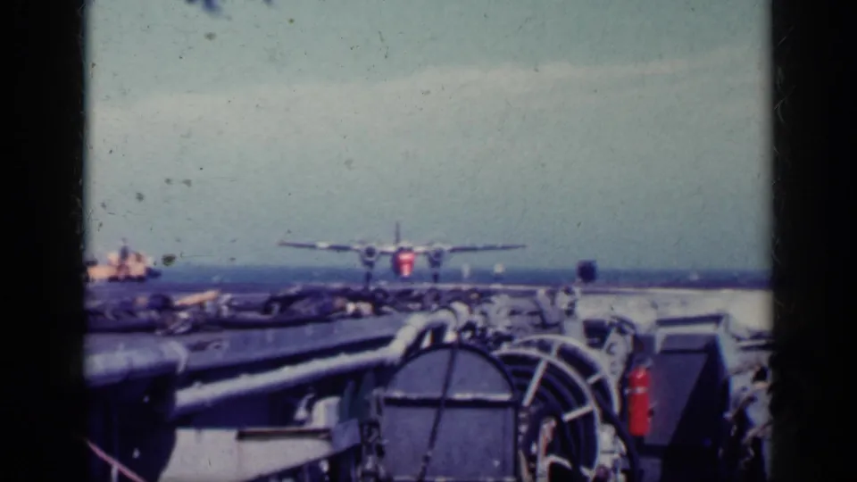 1962 Cuba 8mm frame showing a military plane near the carrier deck, with ship hardware, railings, and ocean horizon visible.
