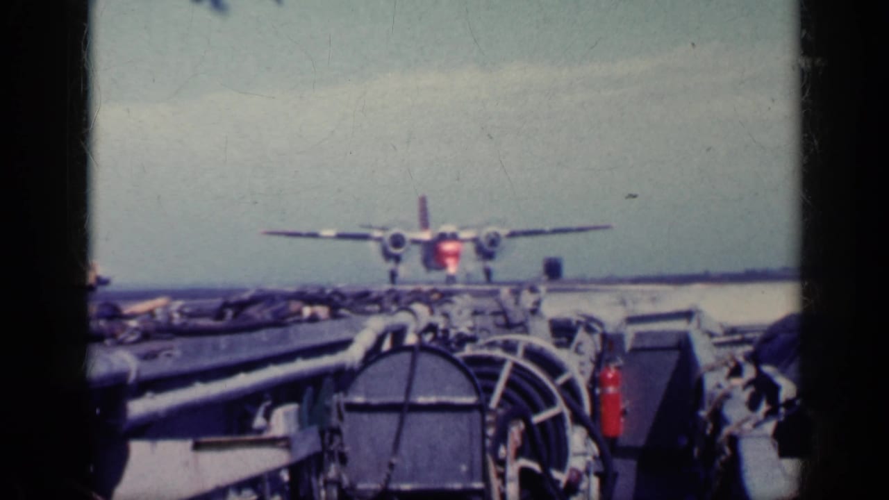 1962 Cuba-labeled 8mm frame showing a twin-engine propeller aircraft aligned on a carrier deck, seen past deck equipment and railings.