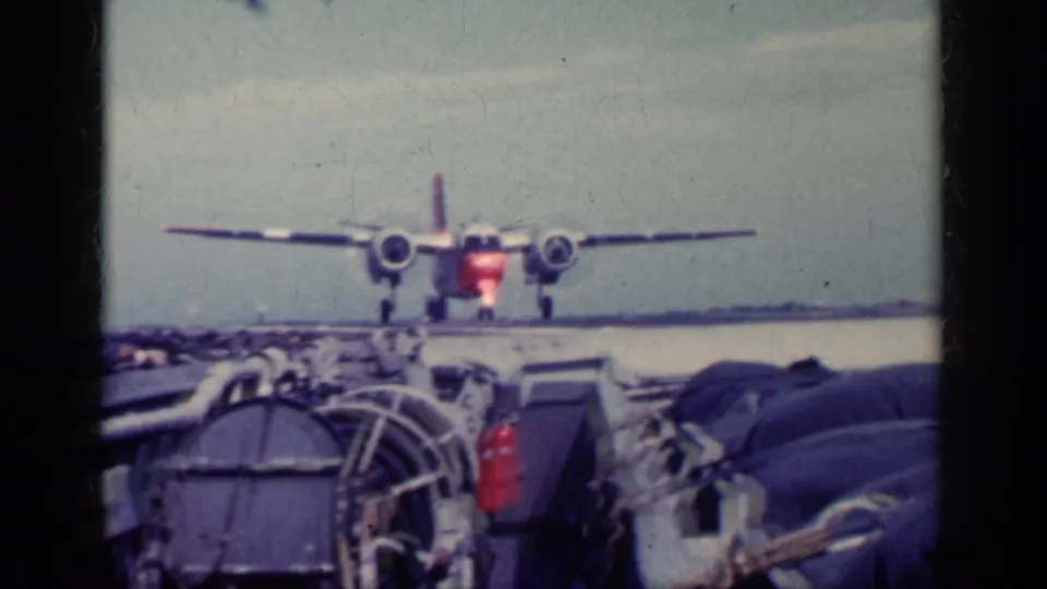 Twin-engine propeller aircraft on a carrier deck in Cuba-labeled 1962 footage, framed by 8mm gate edges and foreground deck gear.