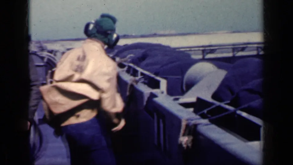 Carrier deck crew member seen from behind in 1962 Cuba-labeled 8mm footage, with deck structures, covered gear and open water beyond.
