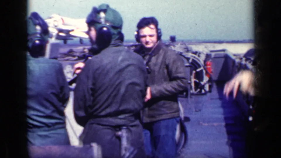 Adult military men in helmets and heavy jackets gather on the ship deck, with a jet aircraft visible in the background.