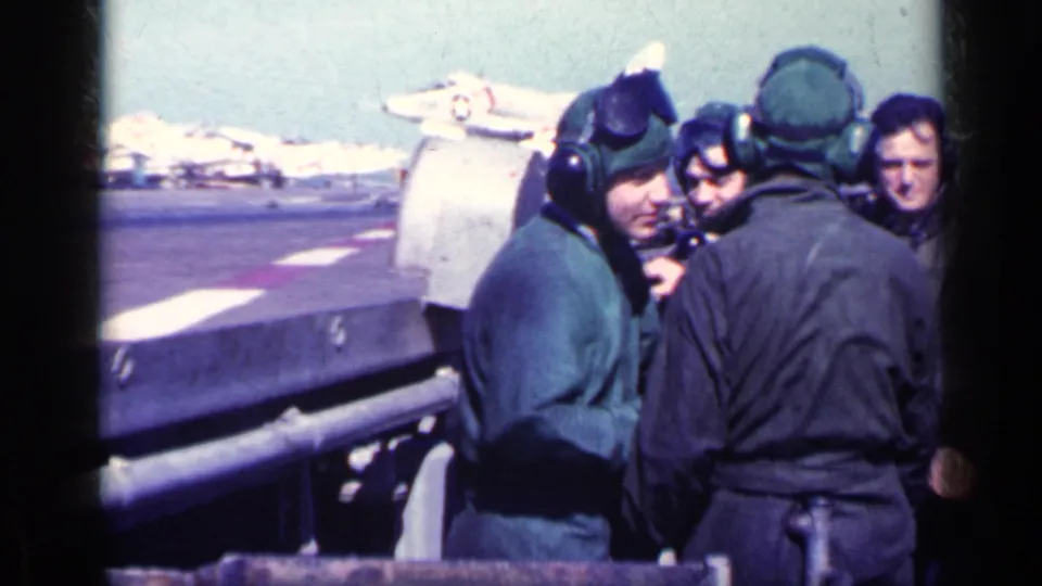 Adult military men in deck clothing gather near a railing on the ship flight deck, with jet aircraft visible beyond them.