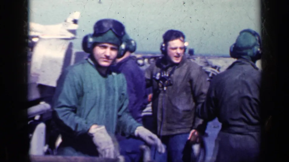 A helmeted adult man wearing goggles and gloves faces the camera on a ship flight deck, with other crew and aircraft behind him.