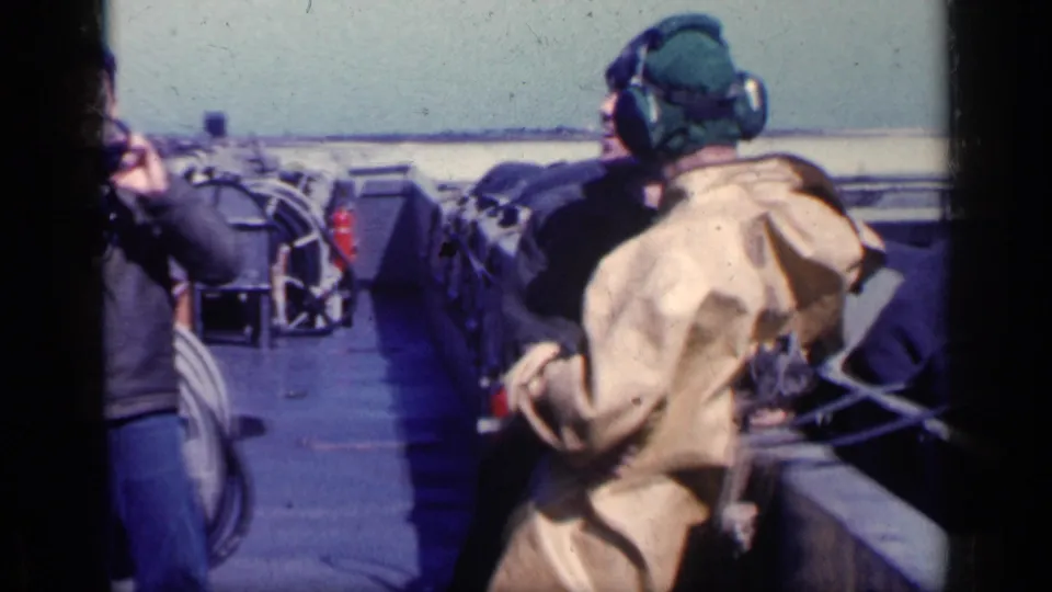 A man in a tan coat and others in flight gear move along a deck lined with equipment in the Cuba 1962 footage.