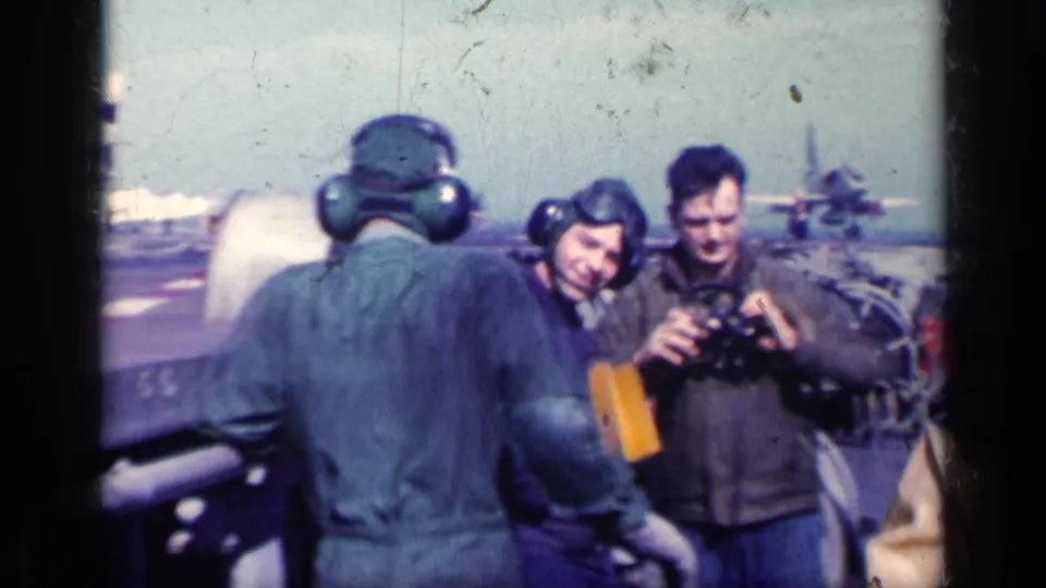 Men wearing ear protection stand on an aircraft carrier deck in Cuba 1962 footage, with a distant jet aircraft and ocean horizon behind them.