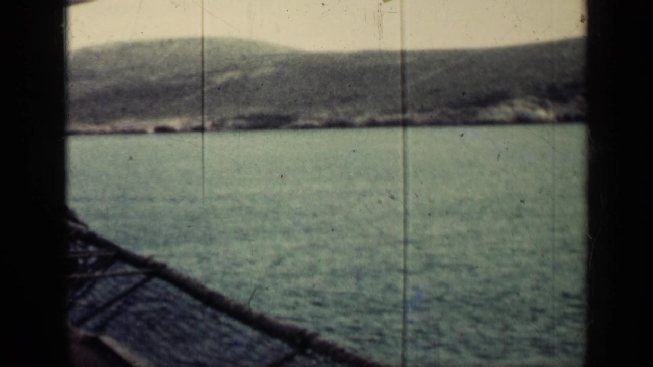 1962 Cuba home-movie view of calm sea and hazy shoreline framed by a dark vertical structure from a moving vessel.