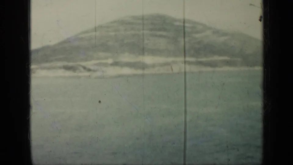1962 Cuba home-movie frame showing calm sea, a pale shoreline, and a rounded mountain under hazy light.