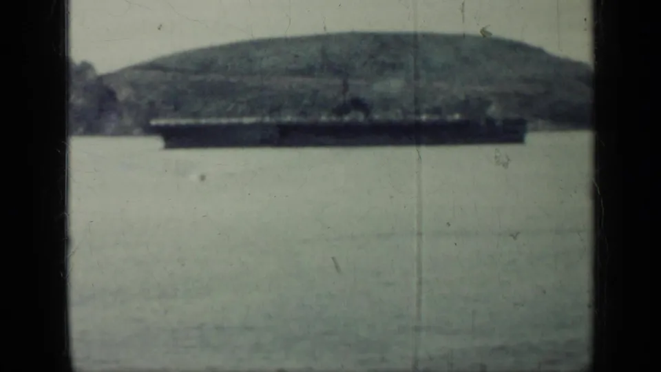 1962 Cuba 8mm frame showing a large dark ship on calm water with a rounded hill and rocky shoreline in the background.